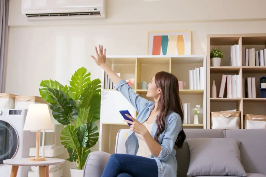 Woman holding smartphone setting remote control for wall-mounted air conditioner in cozy living room.