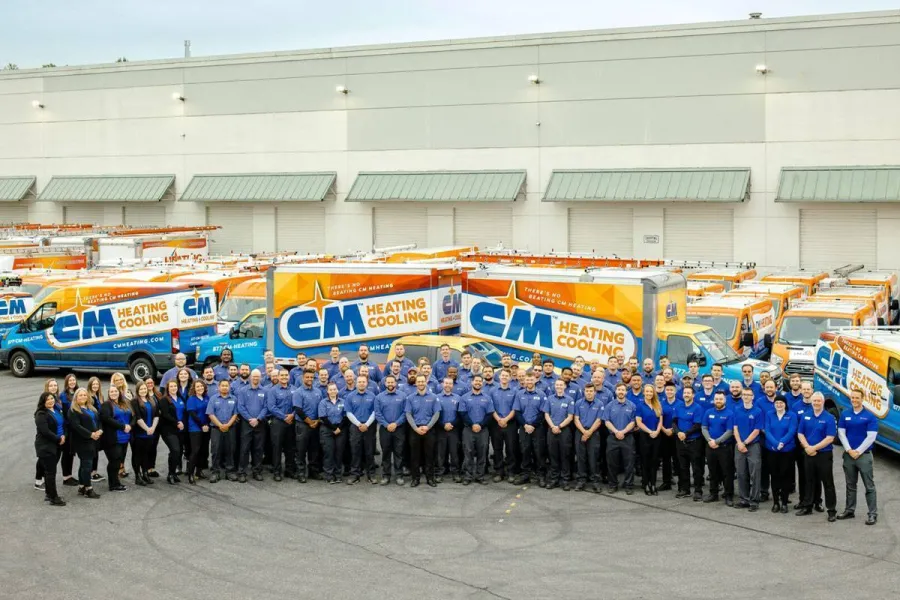 Group photo of CM Heating Cooling employees standing in front of branded company service vans outside a warehouse.