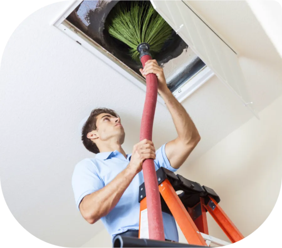Technician cleaning a ceiling air duct using a brush attached to a long hose while standing on a ladder.