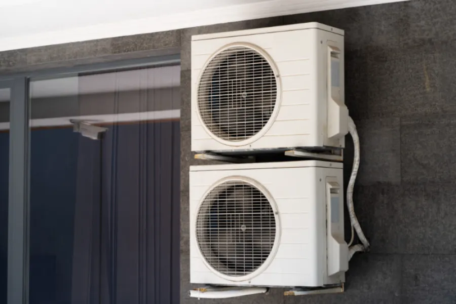 Two white air conditioning units mounted vertically on a dark exterior wall beside a large glass window.