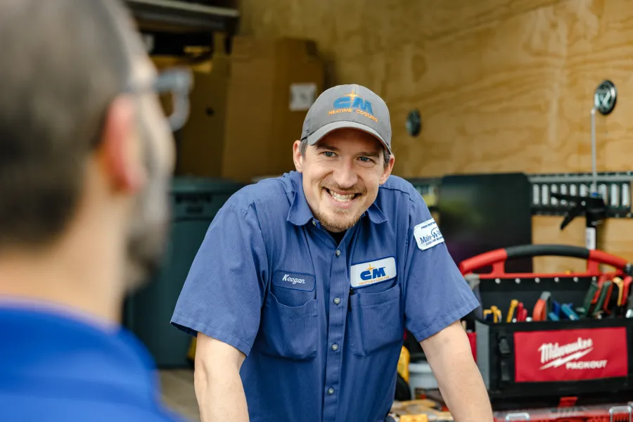 Smiling technician wearing CM Heating and Cooling uniform talking to a customer in a workshop with tools behind.
