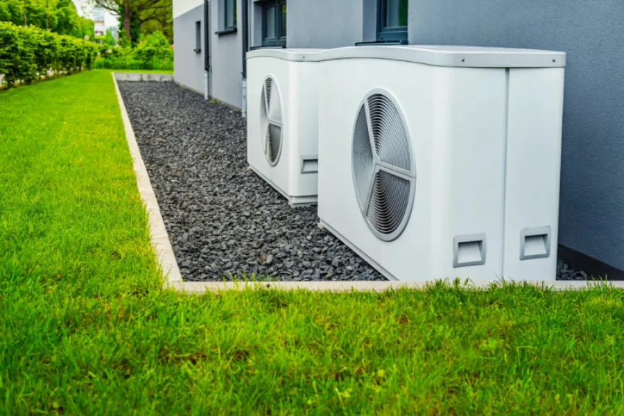 Two modern white air conditioning units installed outside a grey house on gravel next to green grass.