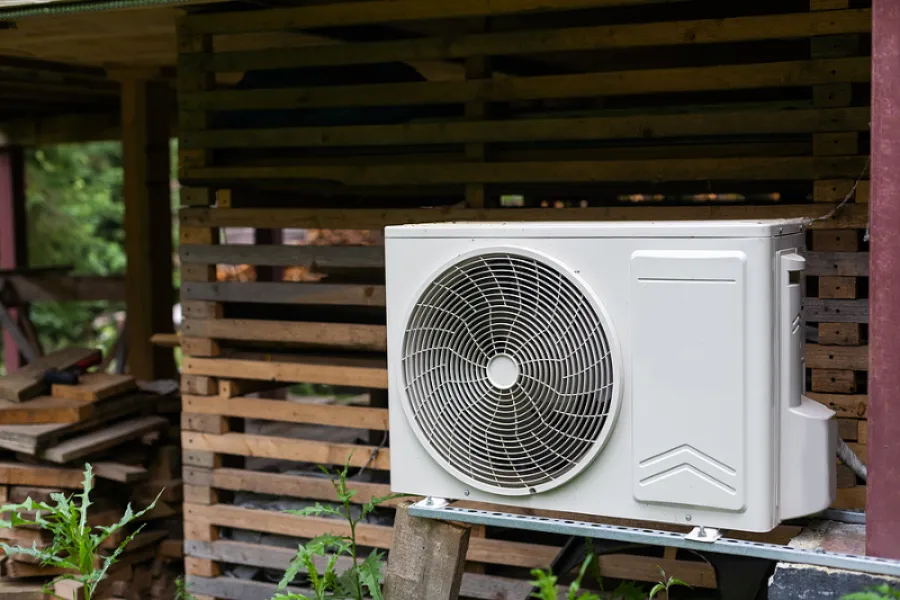 Outdoor white air conditioning unit mounted on metal bracket against wooden wall with greenery nearby.