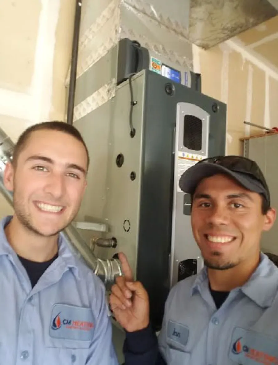 Two HVAC technicians smiling in uniform standing next to a heating unit inside a garage or utility room.