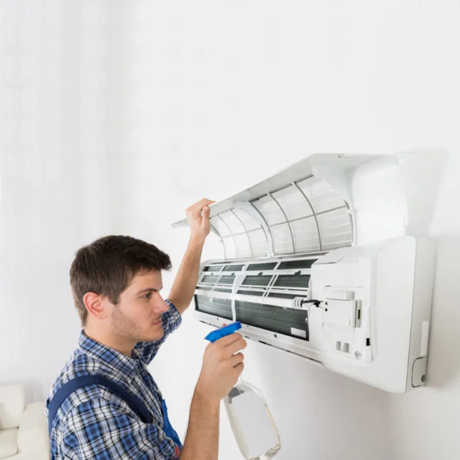 Technician cleaning a wall-mounted air conditioner unit with a spray bottle while inspecting the filter and components.