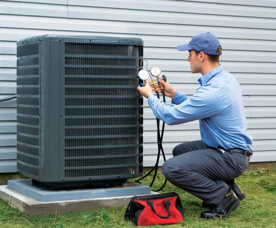 Technician in blue uniform checking outdoor air conditioning unit with pressure gauges near house siding.