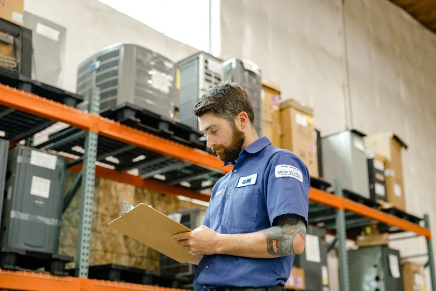 Technician in blue uniform inspecting and taking notes on a clipboard in a warehouse with HVAC units on shelves.