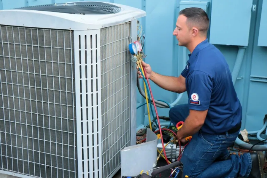 Technician in blue shirt kneels while checking HVAC system with pressure gauges and tools outdoors.