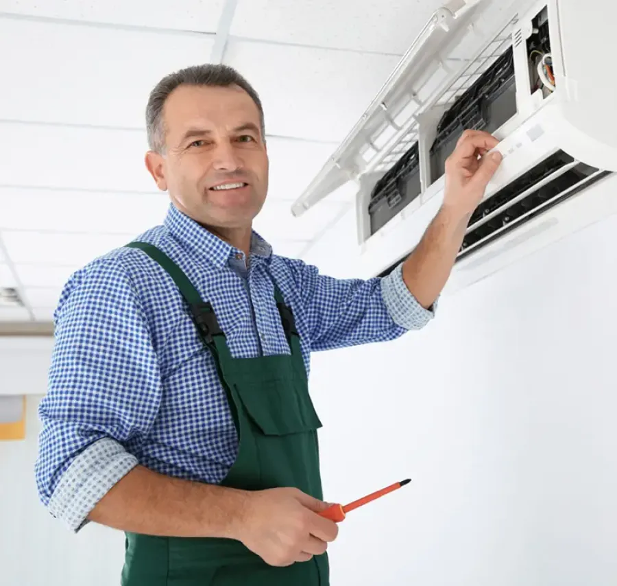 Technician in green overalls repairing or installing a split air conditioning unit on a white wall indoors.