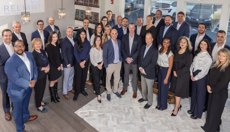 Group portrait of 29 professionally dressed people standing inside a modern office with the Reliant Real Estate sign visible.