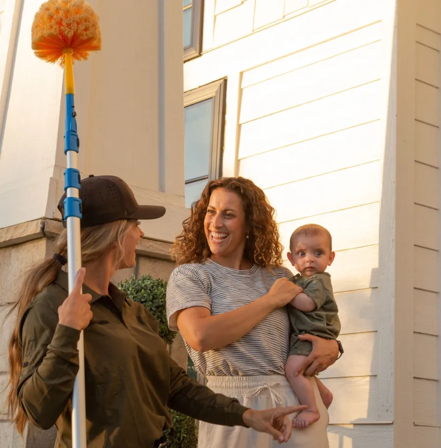 Woman holding baby greets cleaner with duster outside modern home during daytime