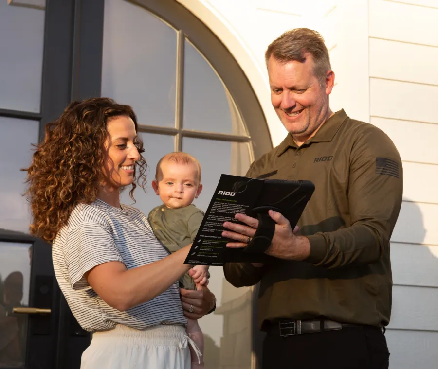 Smiling couple holding baby and reviewing tablet outside home with arched window and white siding