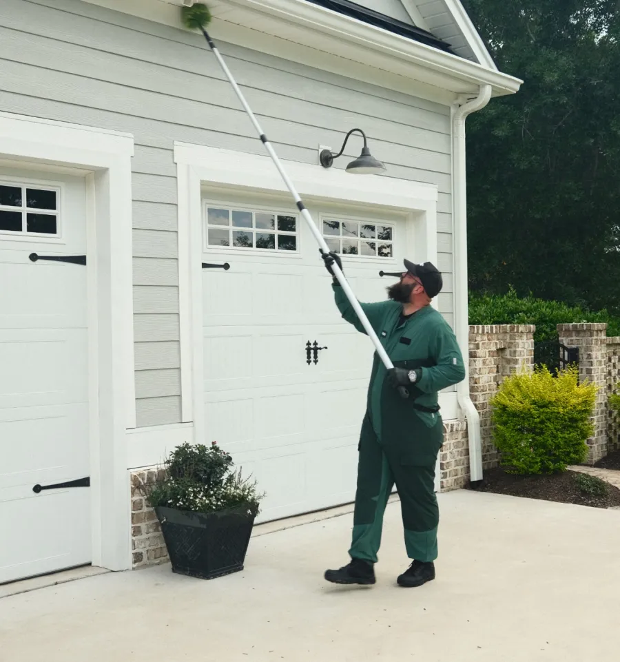 Professional cleaner using an extendable brush to clean outdoor lights on a house exterior near garage doors.