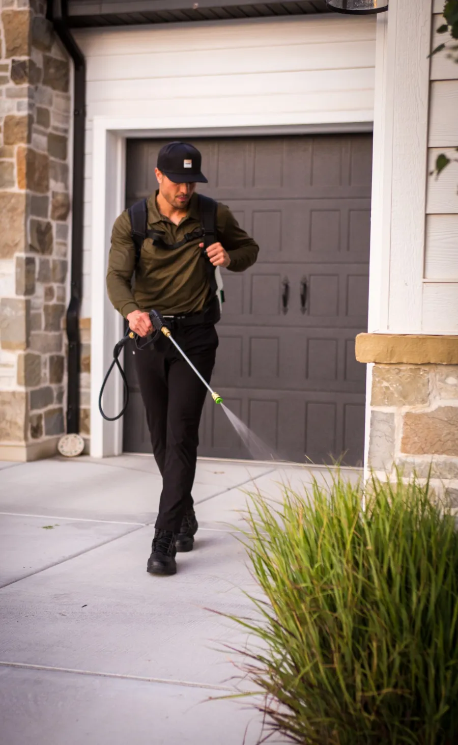 Man wearing casual work clothes spraying pesticide on plants outside a residential garage with stone and wood facade.