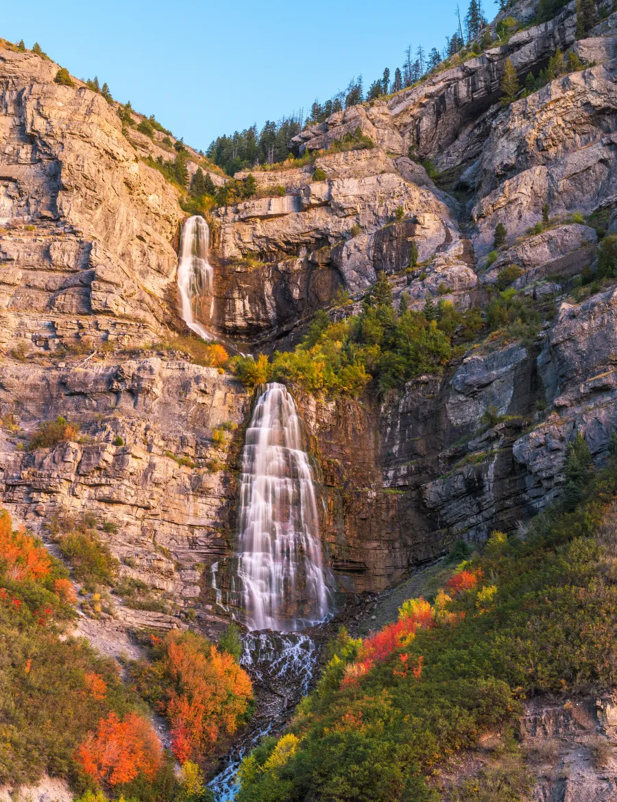 Two-tier waterfall flowing down rocky cliffs surrounded by autumn foliage and green trees under clear blue sky