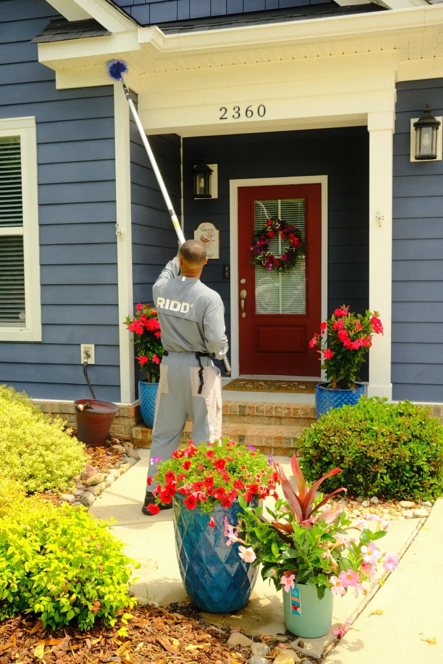 Professional pest control worker in uniform treating exterior of blue house near red door and flower pots.