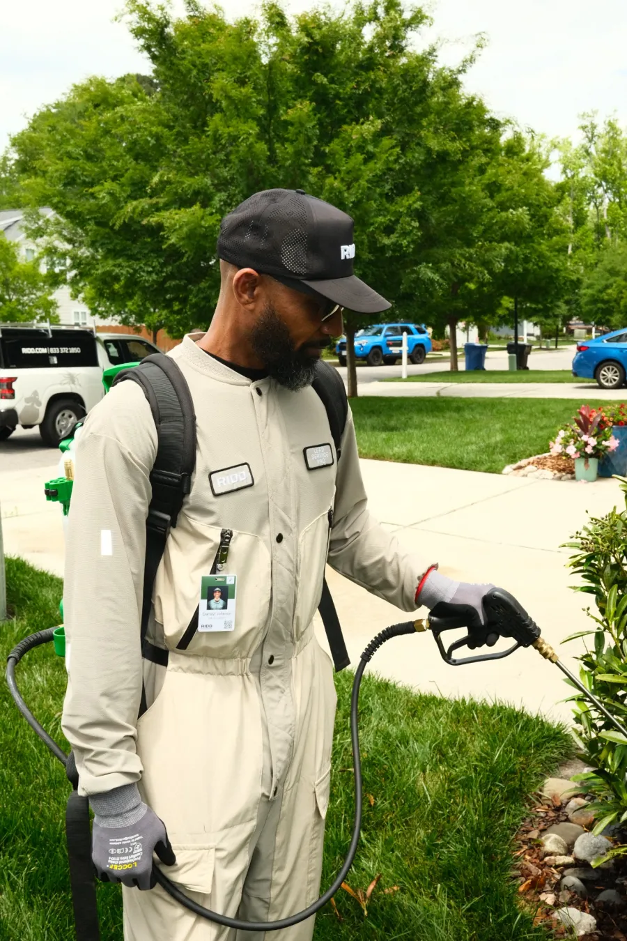 Technician in beige uniform spraying plants outside with equipment and protective gloves near sidewalk and parked vehicles.