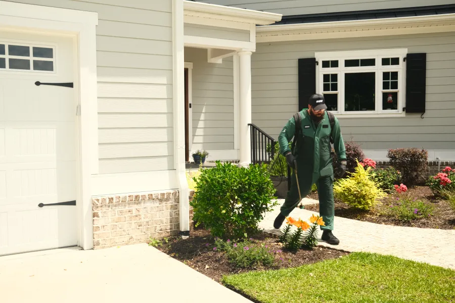 Gardener in green uniform tending flower beds near a white house on a sunny day.