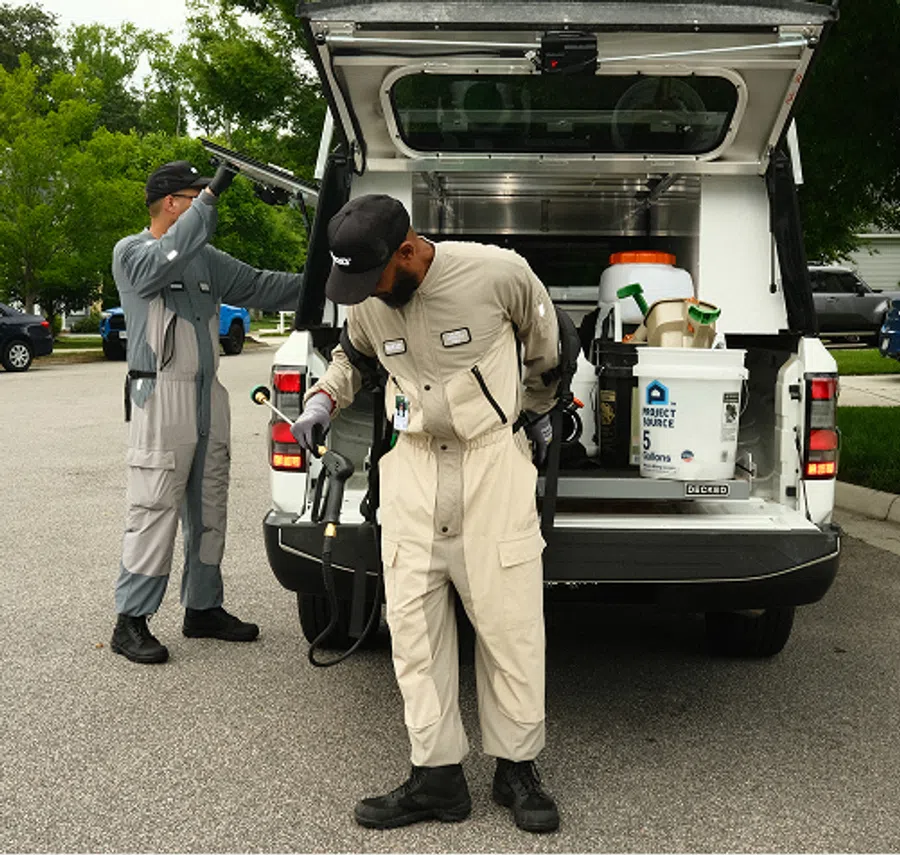Two pest control technicians preparing equipment at the back of a service truck in a residential neighborhood.