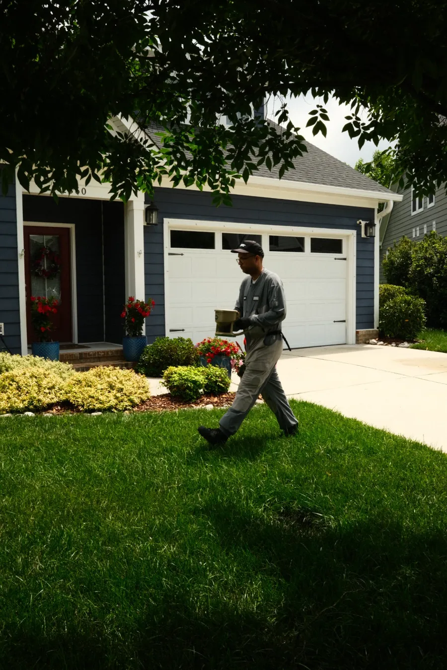 Man in uniform carrying a container walking on grass in front of a blue house with white garage door and flowers.