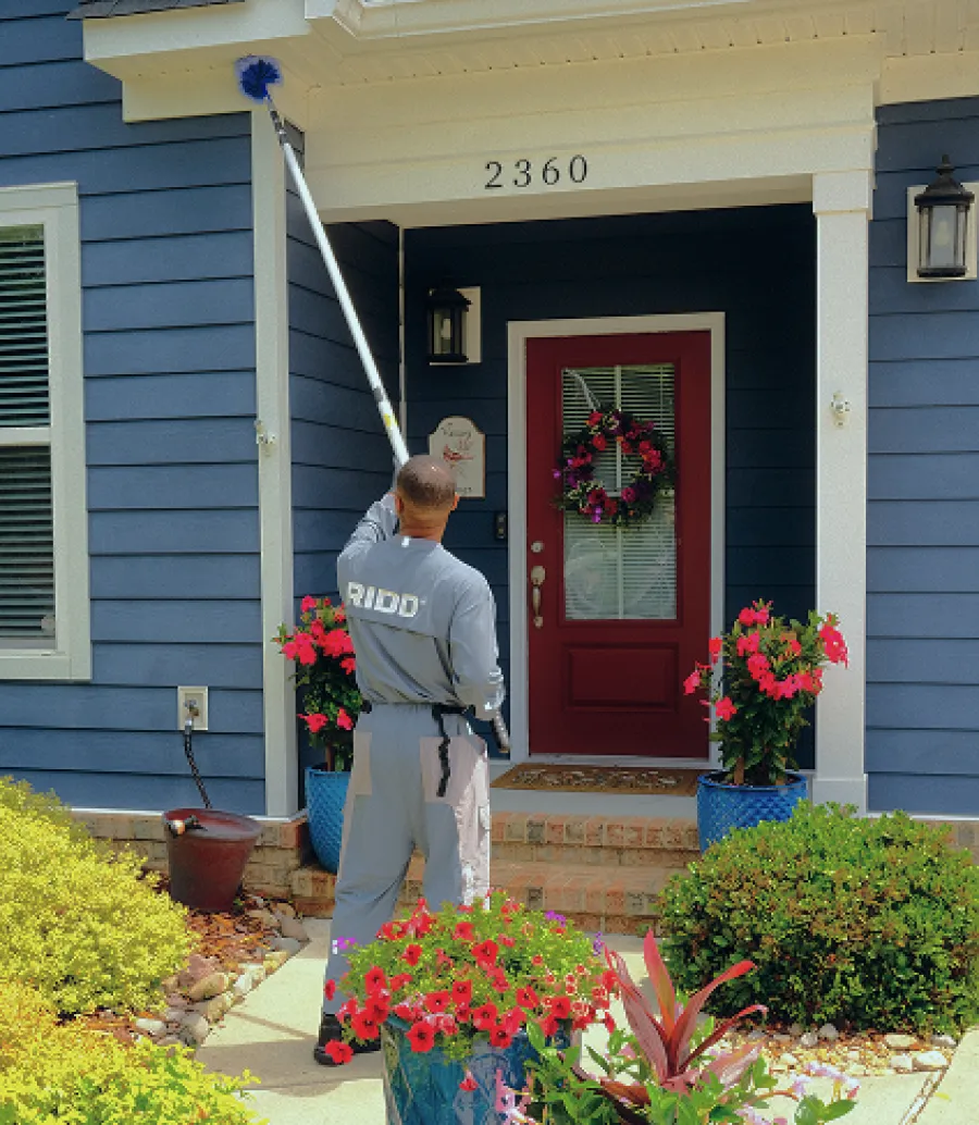 Professional cleaner dusting the exterior near front door of blue house with red door and flower pots