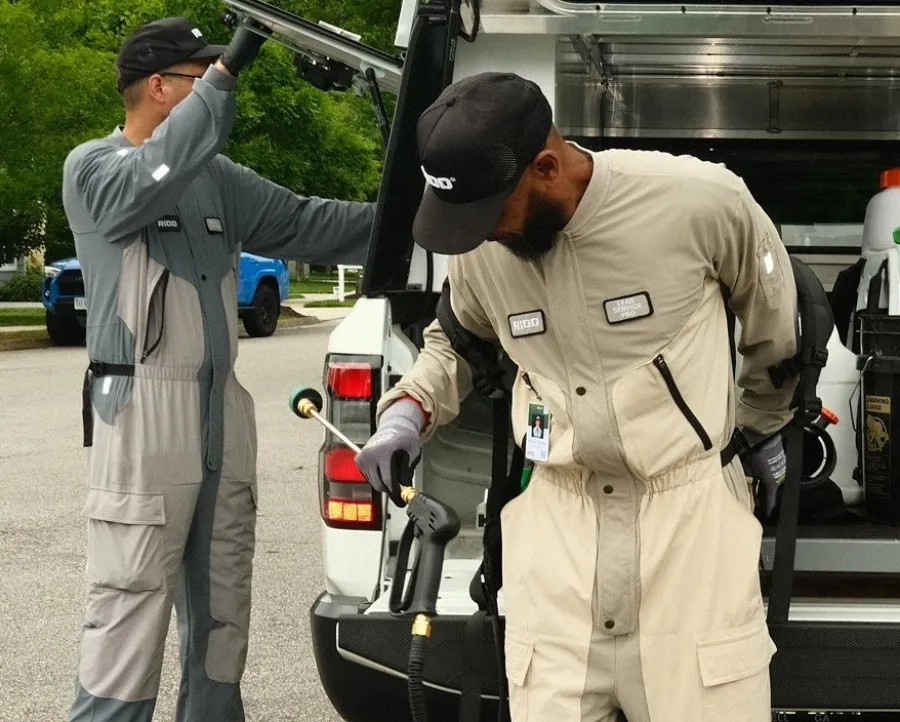 Two technicians in uniform loading tools and equipment into a work van parked on a residential street.