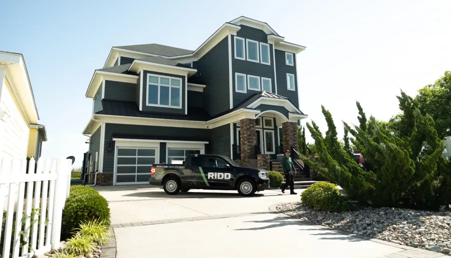 Modern two-story house with dark blue siding, large windows, stone pillars, and a RIDD service truck parked in driveway
