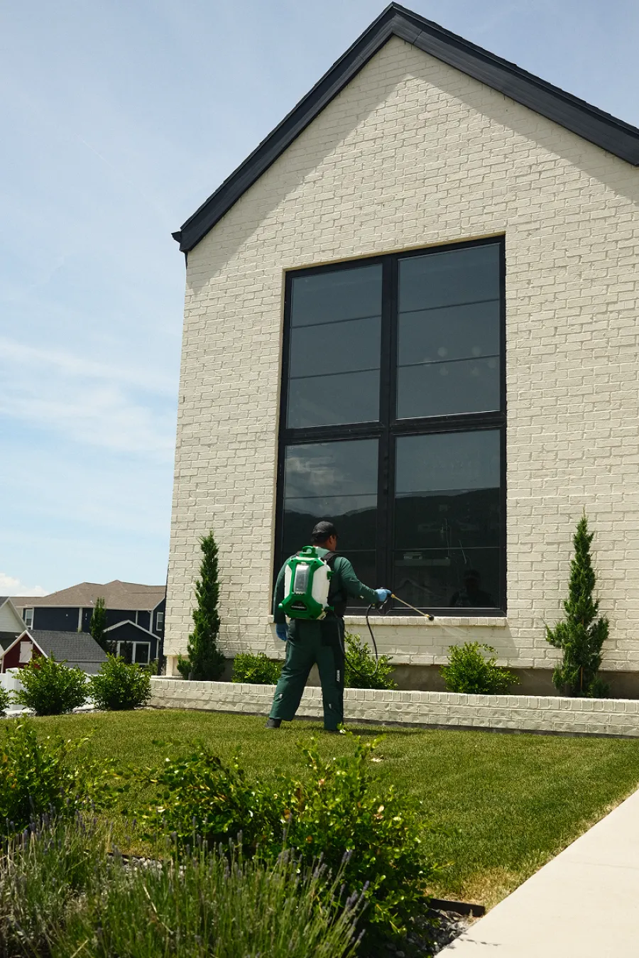Professional using a backpack sprayer to treat plants outside a white brick house on a sunny day