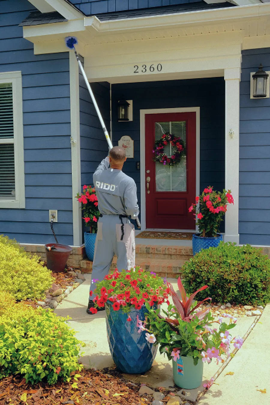 Man cleaning cobwebs from front porch of blue house with red door and colorful flower pots.