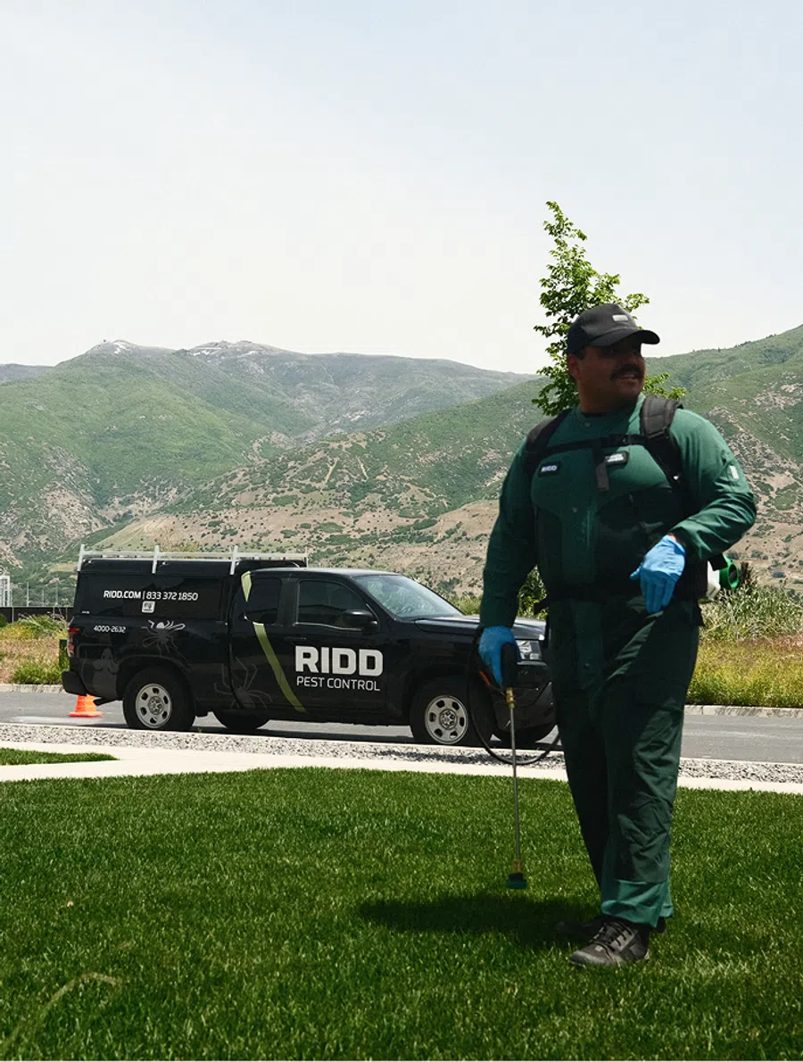 Pest control technician in green uniform with sprayer on lawn, RIDO pest control truck in background mountainous area.