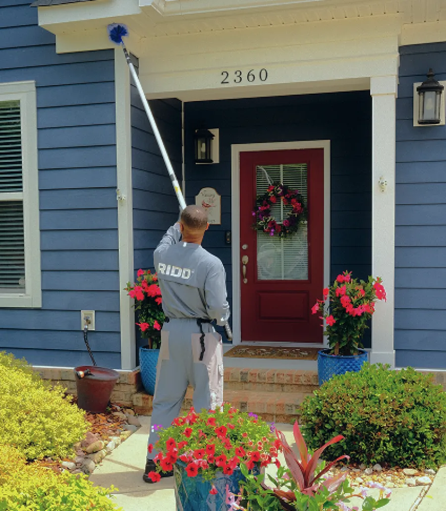 Professional cleaner dusts the exterior of a blue house with red door surrounded by vibrant plants and flowers.
