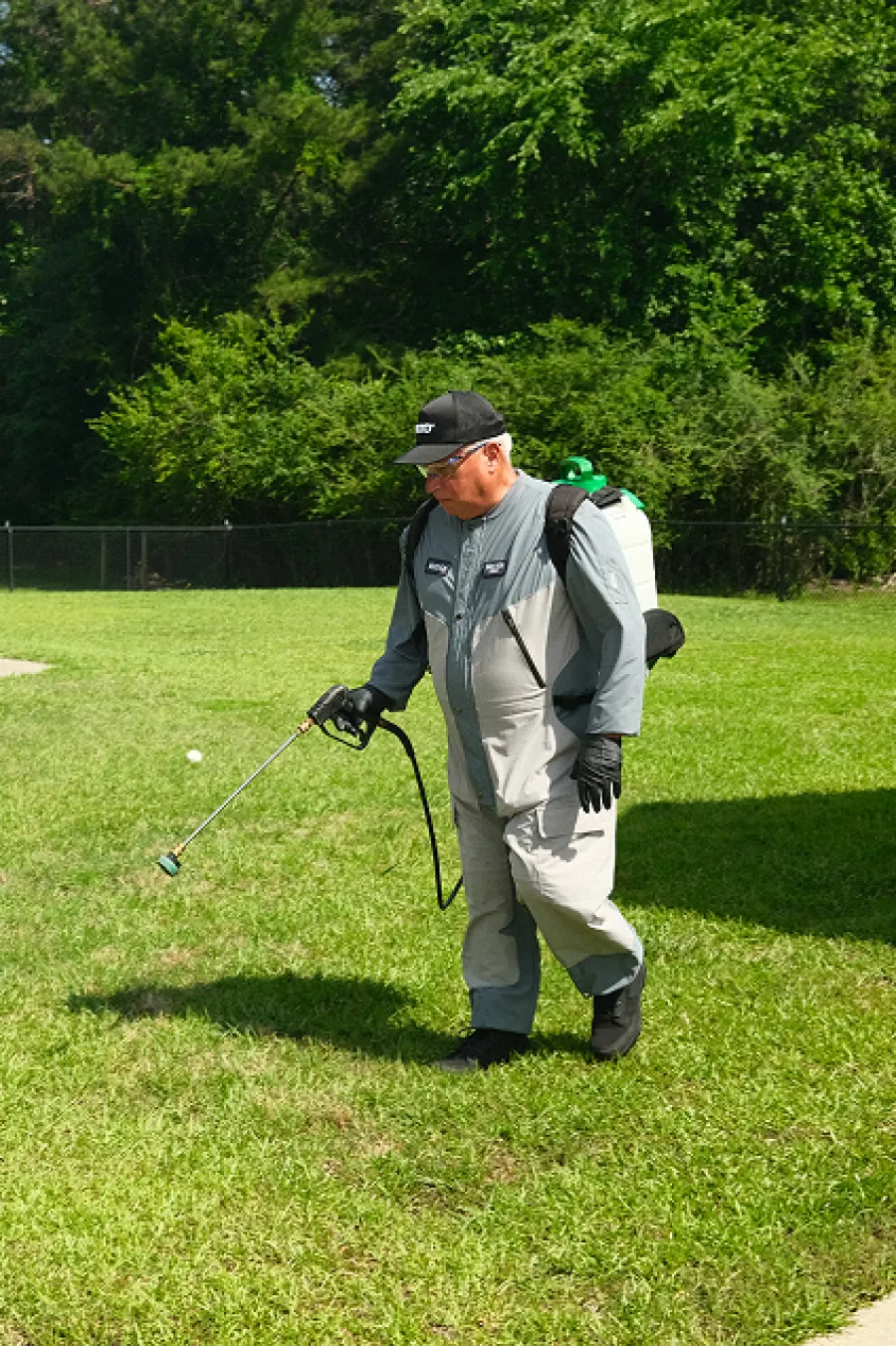 Man in protective gear spraying pesticide on lawn grass with greenery and trees in background.