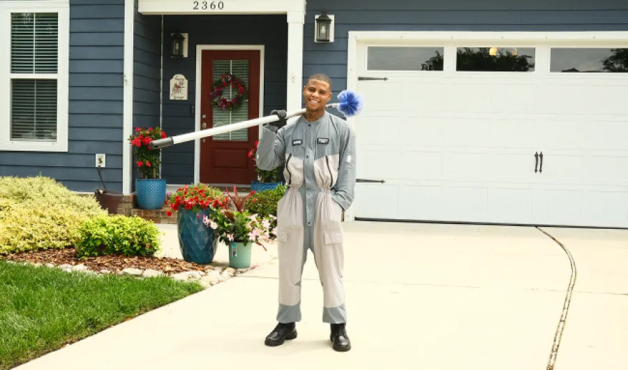 Smiling man in gray work uniform holding cleaning tool stands outside blue house with red door and garage.