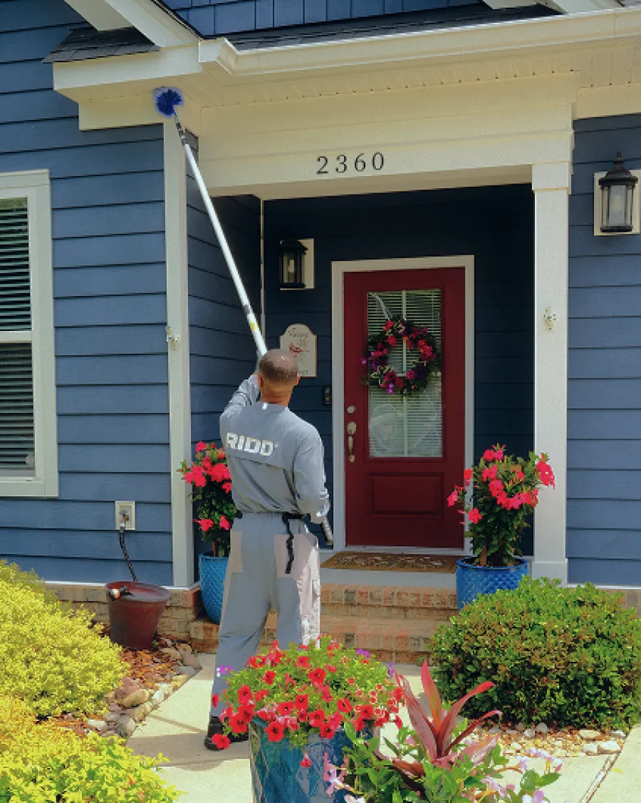 Professional cleaner dusts blue house exterior near red door with flower pots and greenery outside.