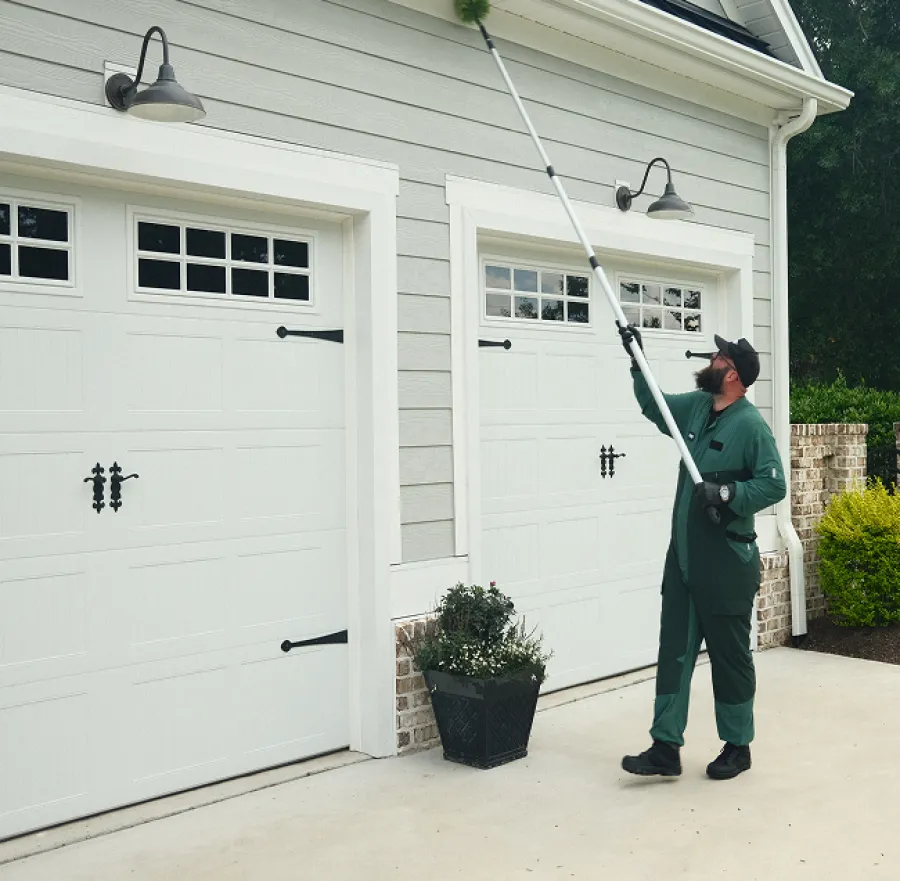 Man using long pole to clean exterior wall light fixtures on gray house with white garage doors.
