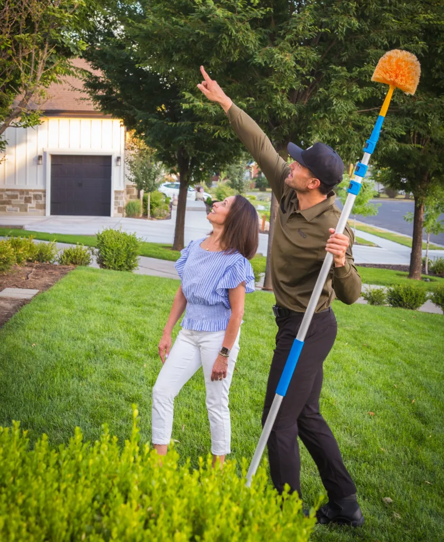 Man holding a long-handled brush points up while woman looks on in a green suburban yard with trees and houses.