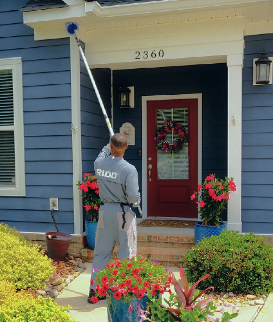 Professional cleaner dusting the exterior near front door of blue house with red door and flower pots