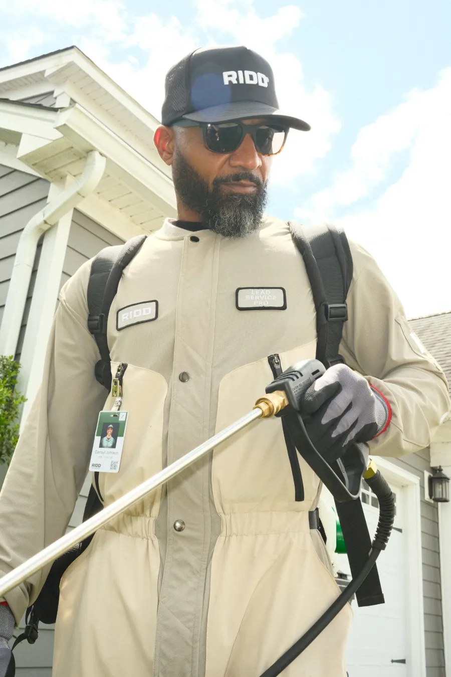 Man wearing protective gear and cap holding a pressure washer wand outside a house on a sunny day