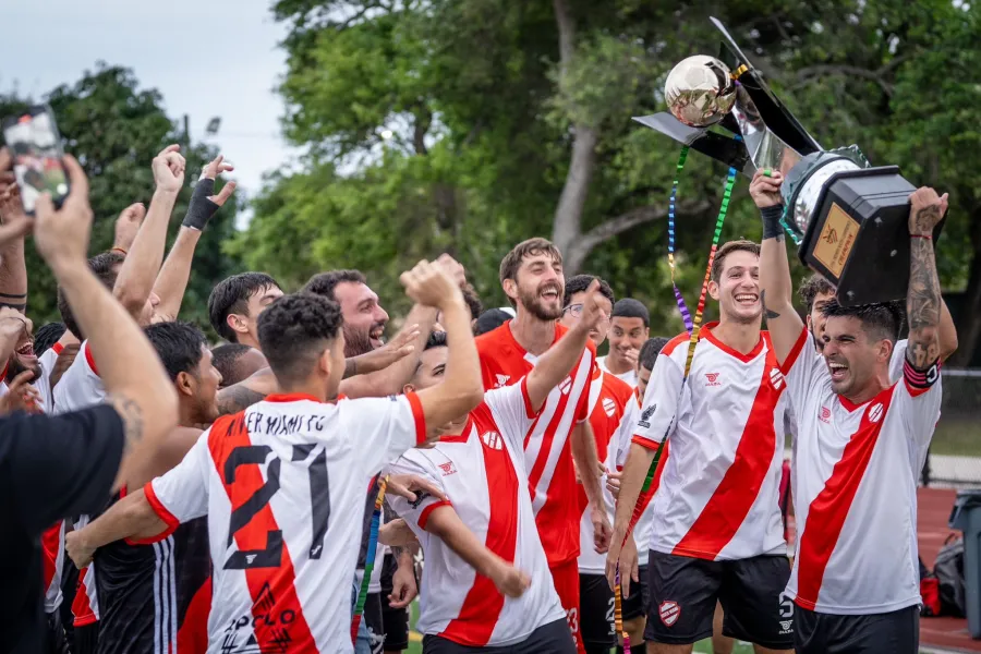Soccer team in white and red uniforms celebrating and lifting a trophy outdoors with joyful expressions.