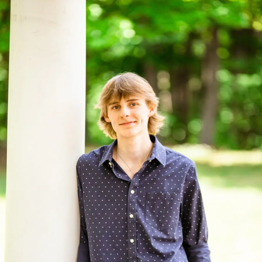 Young man in navy polka dot shirt and white pants leans against a white pillar in a green outdoor setting