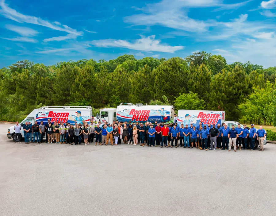 Large group of Rooter Plus plumbing employees posing in front of company trucks outdoors with trees and blue sky background