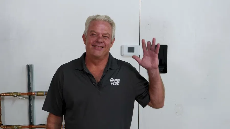 Man in black Rooter Plus shirt waving with a smile in front of a white wall with pipes and a thermostat.