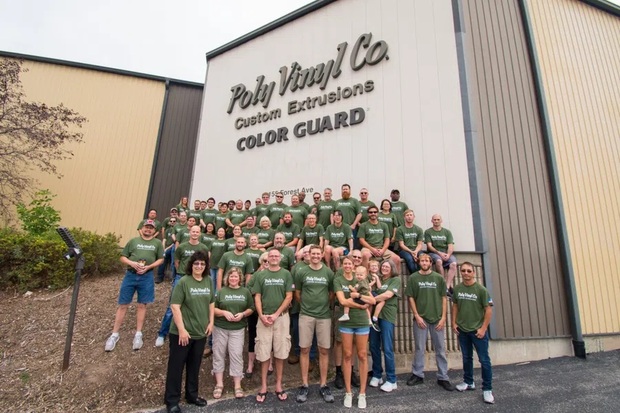 Group of Poly Vinyl Co. employees wearing green shirts posing outside company building under company sign.