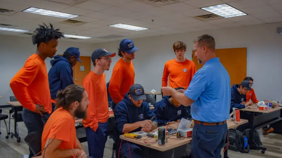 Instructor demonstrating electronics assembly to a group of students in orange uniforms in a classroom setting.
