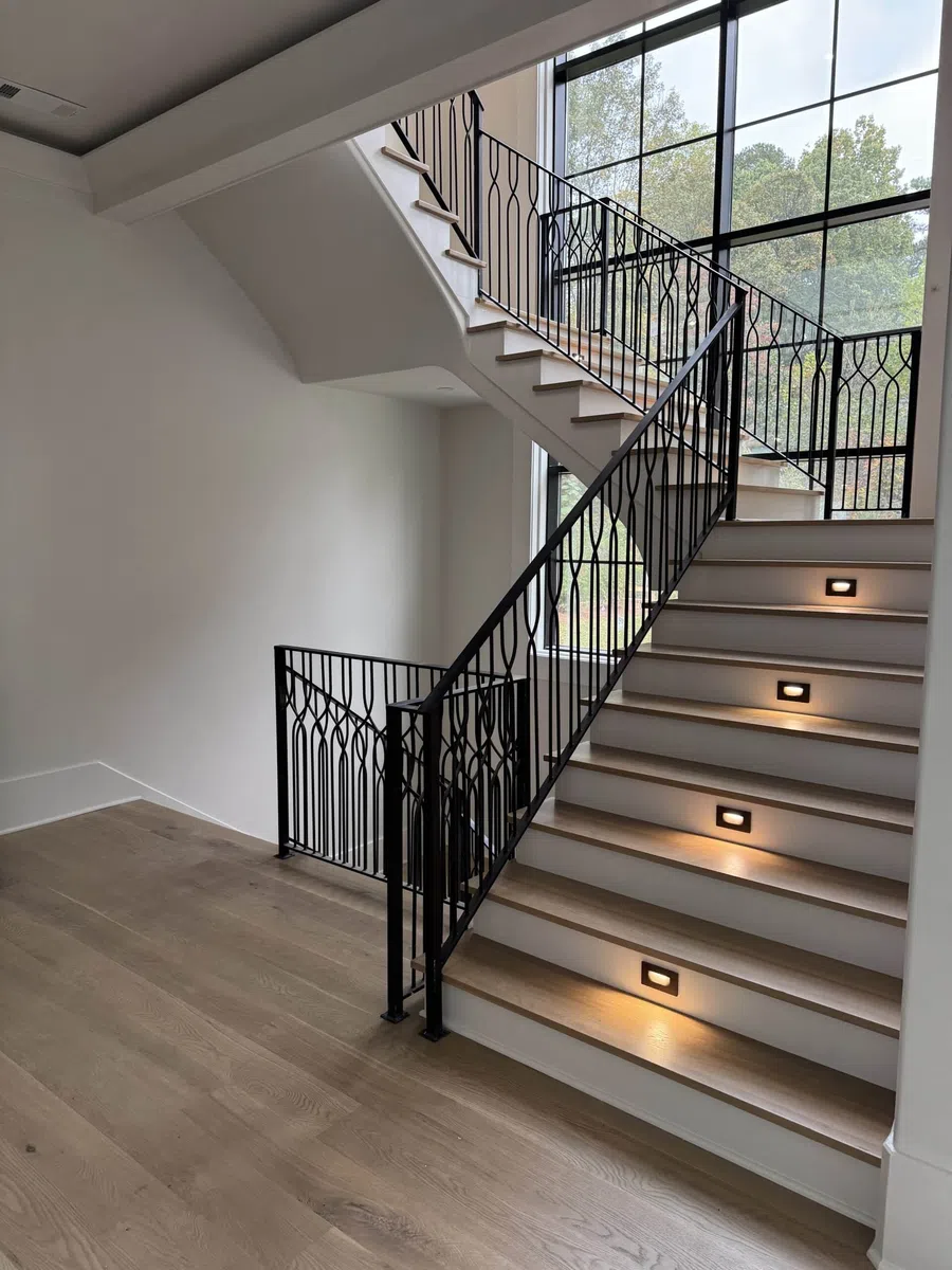 Modern staircase with wooden steps, black wrought iron railing, and built-in step lighting by large window.