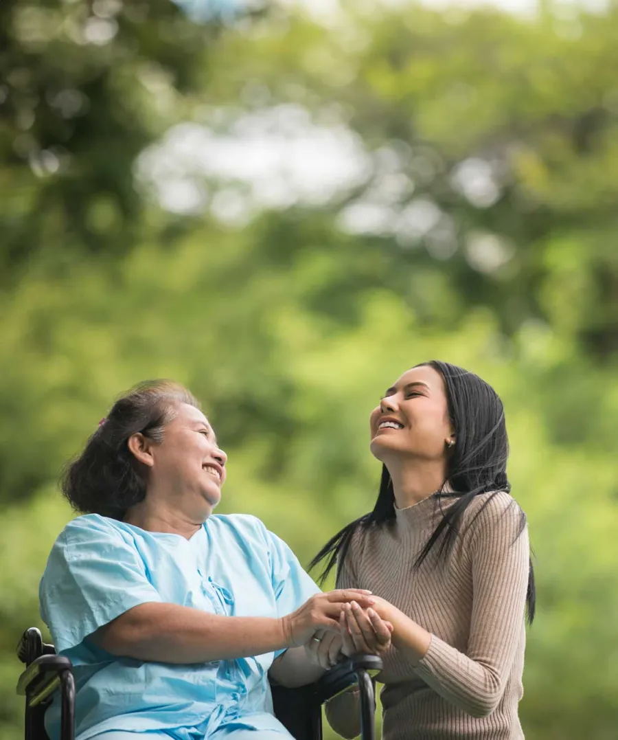 Older woman in wheelchair and younger woman holding hands and smiling outdoors with green trees in background