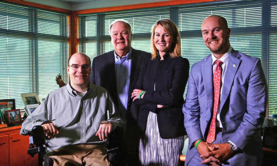 Four professionals smiling in an office, one seated in a wheelchair, two men and one woman standing.