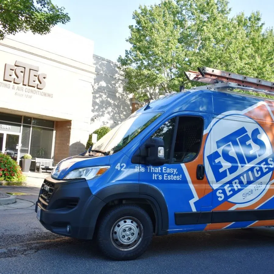 Estes Services blue and orange service van parked outside Estes Heating & Air Conditioning office with technician standing nearby.