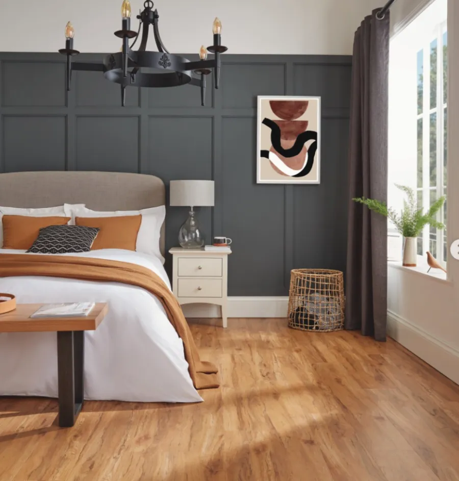 Cozy modern bedroom with dark gray paneled wall, white bed, rust throw blanket, and wood flooring.