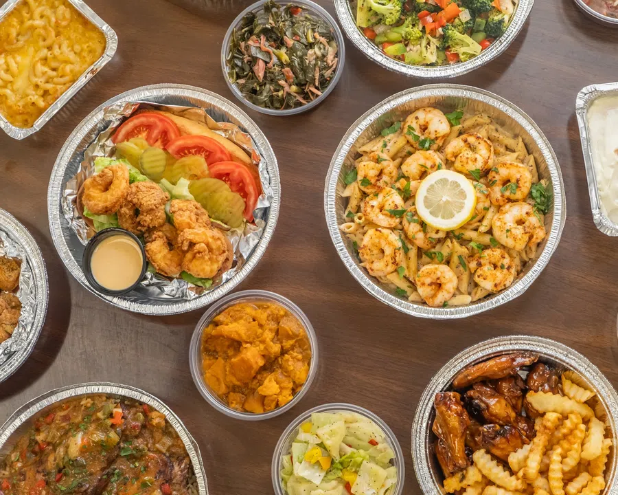 Takeout meal trays with fried shrimp sandwich, shrimp pasta, crinkle fries, mac and cheese, and assorted sides on wooden table.